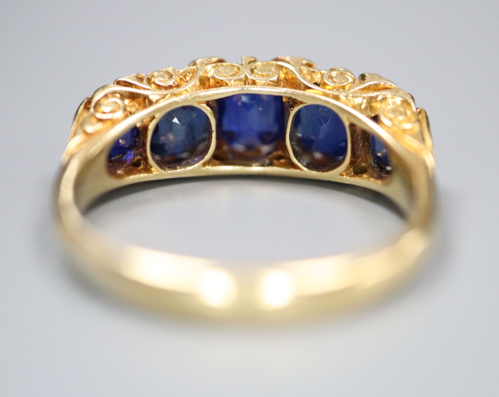 A George V 18ct gold and graduated five stone sapphire set half hoop ring, with diamond chip spacers,